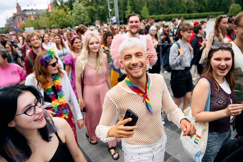 A young man with rainbow makeup, white hair, a mesh shirt and white jeans smiles and walks among a crowd of Pride parade marchers