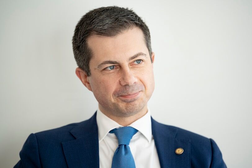 Pete Buttigieg is a white cleanshaveb brunette man of 42 years of age. In this image he sits in a white room while wearing a blue blazer, white shirt and light blue tie while lightly smiling, his eyes looking off to the right.