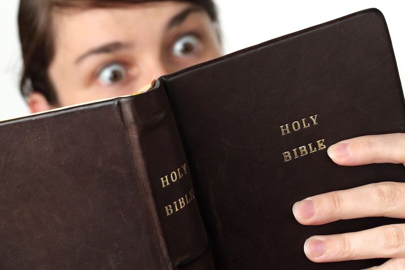 A white-skinned, brown-haired person's eyes open wide in surprise as they read a Holy Bible