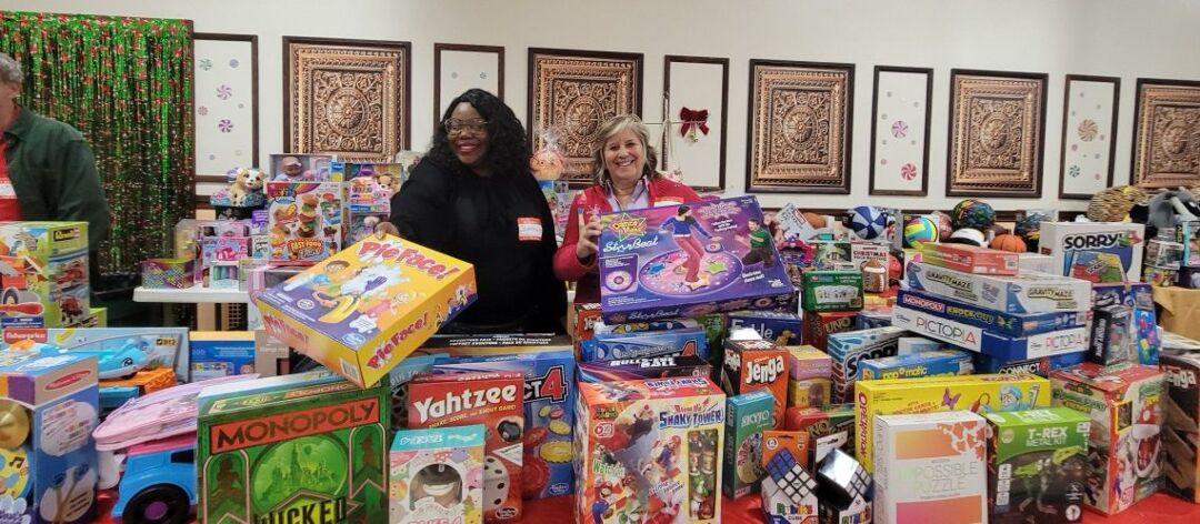 A Black woman and white woman stand in front of a table filled with various board games and toys.