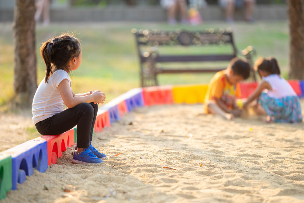 Little girl sitting lonely watching friends play at the playground.