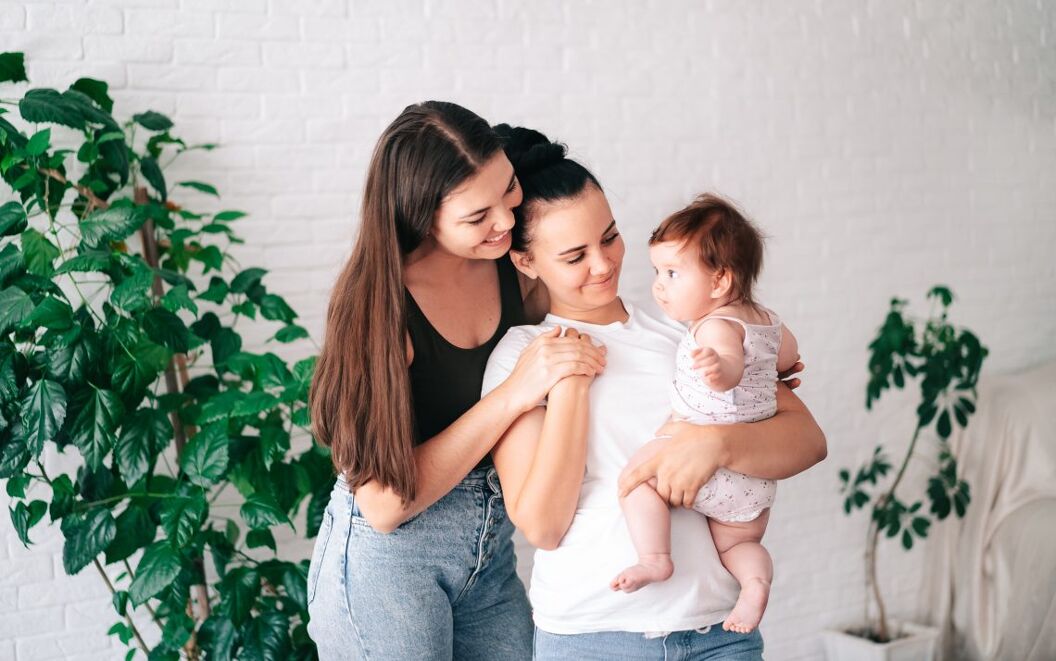Two beautiful young women in casual clothes hug and look at their little child in the apartment. the concept of lgbt people. lesbian marriage and adoption, homosexuals-a lesbian couple.
