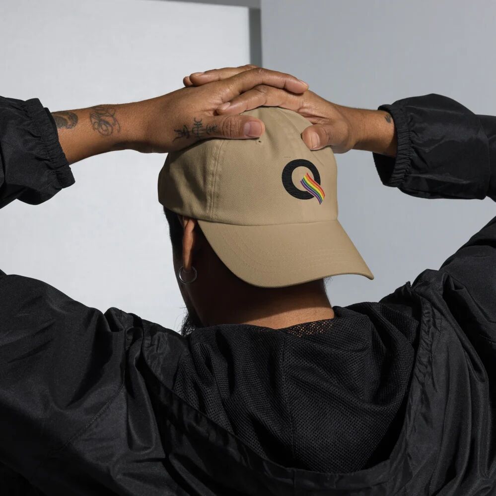 A person wearing a black track jacket and the rainbow Q embroidered dad cap in beige backwards on their head with their hands resting on top